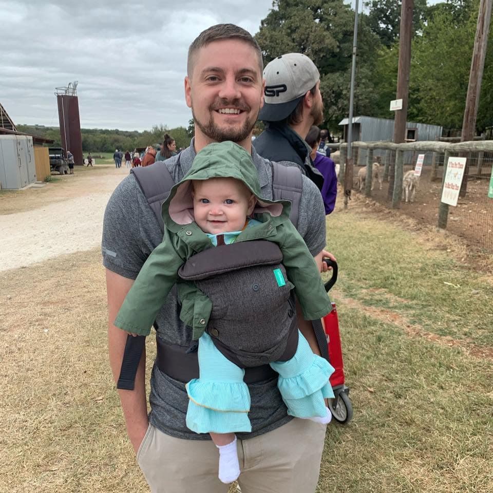 Smiling man carries a baby in a green hooded jacket using a front-facing baby carrier.